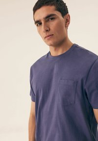 Fioletowa bawełniana koszulka z krótkim rękawem, z kieszonką na piersi, okrągłym dekoltem oraz subtelnym, teksturowanym wzorem na materiale.