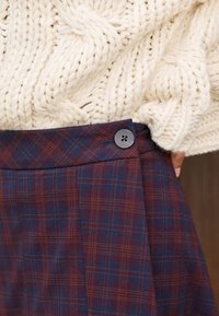 Volnene karo hlače v mornarsko modri in bordo barvi z subtilnimi rdečimi črtami, z stranskim zapiranjem na gumbe in čistimi, krojenimi gubami.