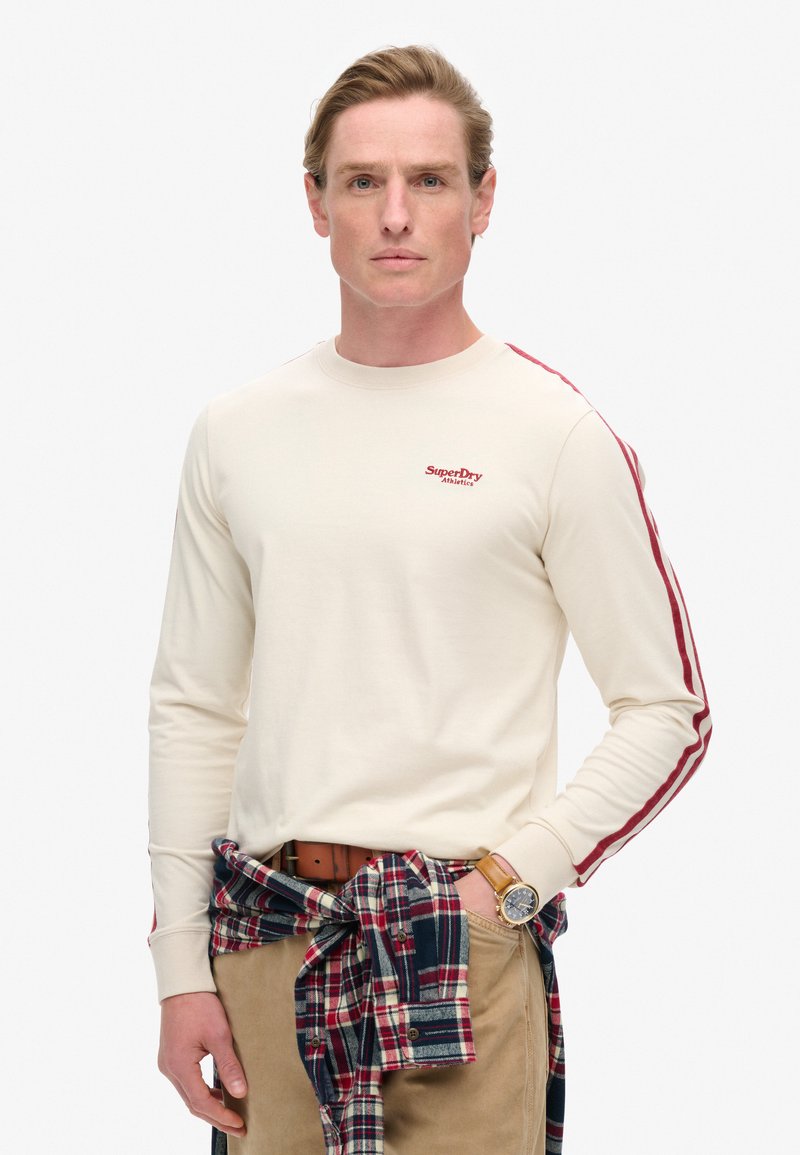 Crèmekleurig overhemd met lange mouwen en rode zijstrepen en geborduurd "SuperDry" logo. Draagt een geruit overhemd geknoopt om de taille. Beige broek en horloge zichtbaar.