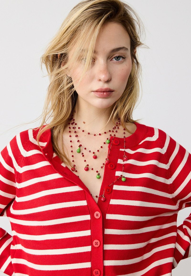 Jeune femme aux cheveux blonds portant un cardigan rayé rouge et blanc et un collier de perles rouge avec des breloques vertes et rouges.