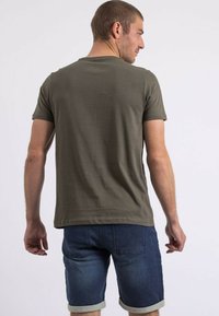 T-shirt en coton vert olive à manches courtes, vue de dos, associé à un short en denim bleu foncé avec des ourlets roulés et des poches arrière.