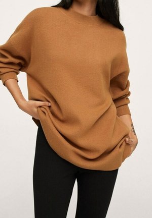 Person in schwarzen Hosen trägt einen lockeren, camel-farbenen Pullover mit langen Ärmeln, gerippten Bündchen und Saum, die Hände auf den Hüften ruhend.