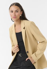 Blazer beige con un design sartorial, bottoni neri e chiusura a un petto, abbinato a un top nero e pantaloni a vita alta.