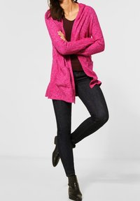 Rosa gestrickte Strickjacke mit Kapuze, offenem Vorderteil und zwei Taschen, darüber getragen ein dunkles Oberteil, kombiniert mit dunklen Jeans und Stiefeletten.