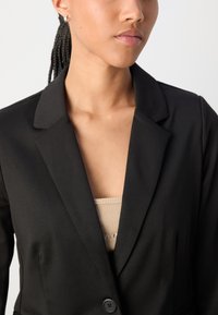 Svart blazer med notched lapels, enkel knappstängning och slät tyg. Bärs över en beige topp med en enkel textur.
