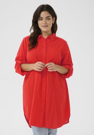 Chemise rouge à boutons avec un col rigide, des manches retroussées, un tissu texturé et une poche, présentant un ourlet plus long et arrondi.