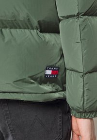Doudoune verte avec motif matelassé horizontal, structure compacte et poignets élastiques. Comprend un patch logo TOMMY JEANS sur le côté.