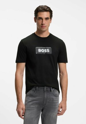 Mand med mørkt hår iført sort t-shirt med sløret "BOSS" tekst og falmede grå jeans, der står mod en hvid baggrund.