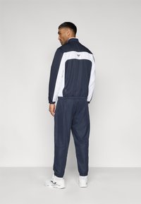 Tumši zils un balts sporta jaka ar elastīgiem aprocēm un jostasvietu, komplektā ar atbilstošām biksēm; uz muguras redzams logotips un brīvs piegriezums.