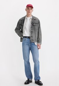 Denim jacket in gray with front pockets, white t-shirt, striped shirt underneath, blue straight-leg jeans, and black shoes. Red cap with logo.