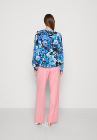 Blusa floreale in un motivo blu, rosa e nero, con maniche lunghe e polsini elastici, abbinata a pantaloni larghi rosa chiaro e tacchi bianchi.