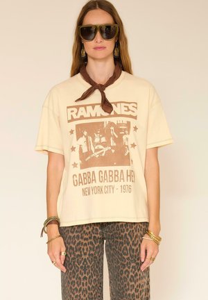 Femme portant des lunettes de soleil surdimensionnées, un t-shirt du groupe Ramones, un foulard marron autour du cou, un pantalon à imprimé léopard, ainsi que plusieurs bracelets et bagues en or.