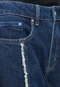 Jeans de denim azul com bainha desfiada, costuras em contraste e um design clássico de cinco bolsos com detalhe em botão de metal.