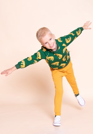 Młody blond chłopiec balansujący na jednej nodze z wyciągniętymi ramionami, ubrany w zielony sweter z nadrukiem tygrysa i musztardowo-żółte spodnie.
