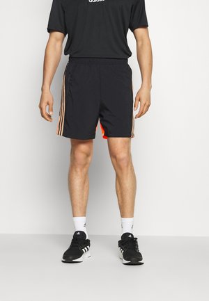 Mężczyzna w czarnych szortach sportowych z pomarańczowymi paskami i czarnych butach Adidas stoi na jednolitym tle.