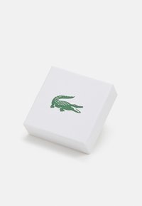 Bijela teksturirana kutija s dizajnom zelenog krokodila na vrhu. Kutija ima čiste linije i minimalistički oblik.