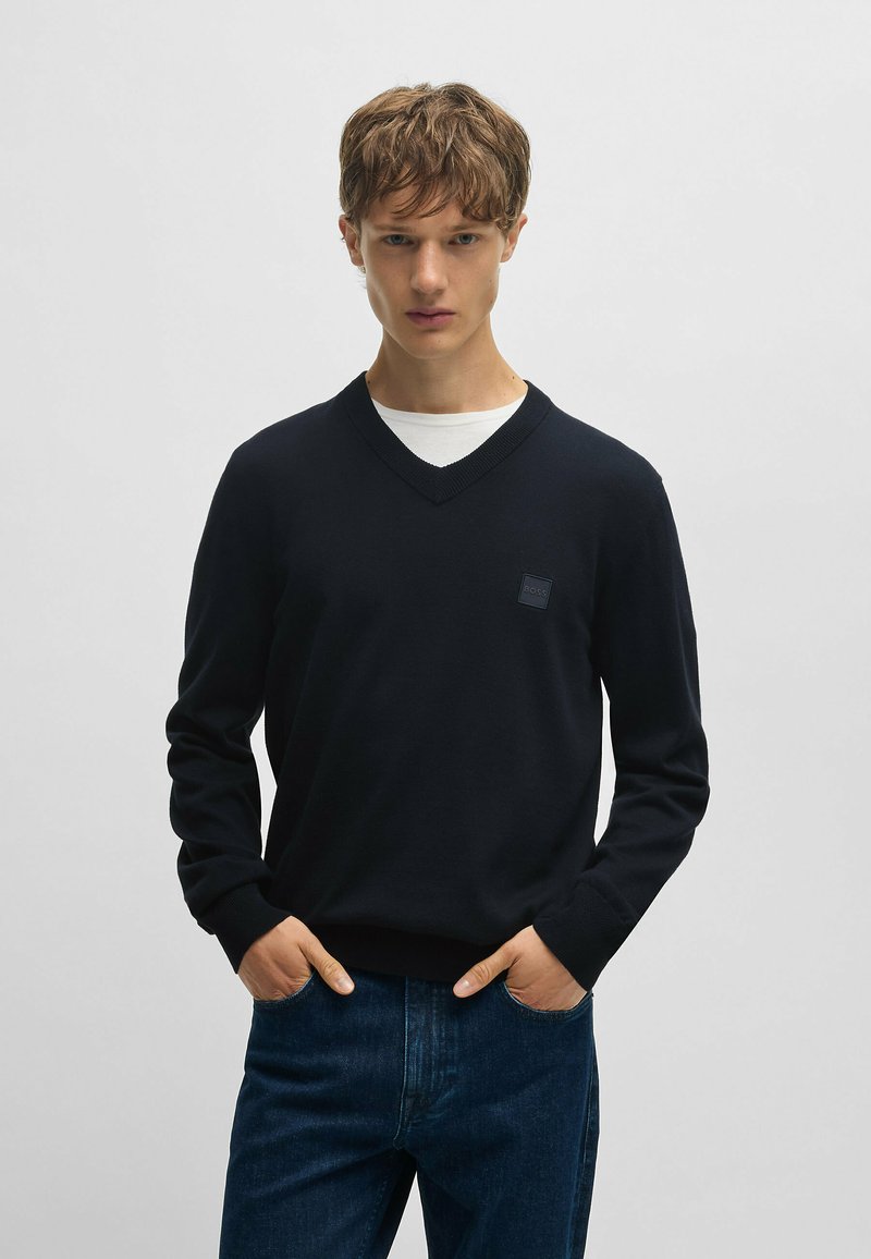 Mörkblå v-ringad tröja i mjukt tyg, med långa ärmar och en liten logotyp på vänster bröst. Matchas med mörka denimjeans.