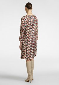 Kleid mit geometrischem Muster in roten, blauen und beigen Tönen. Lange Ärmel und knielanger Schnitt. Gestylt mit beigen Stiefeln bis zum Knie.