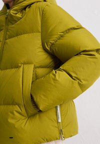 Grüne Puffjacke mit Kapuze, ausgestattet mit einer glatten, glänzenden Textur, Reißverschlusstaschen und elastischen Bündchen. Etikett an der Seitennaht sichtbar.