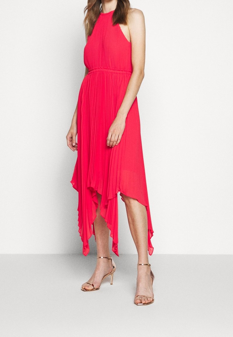 Vestido plisado fucsia con cuello alto, dobladillo asimétrico fluido y textura de satén, combinado con sandalias de tacón desnudas.