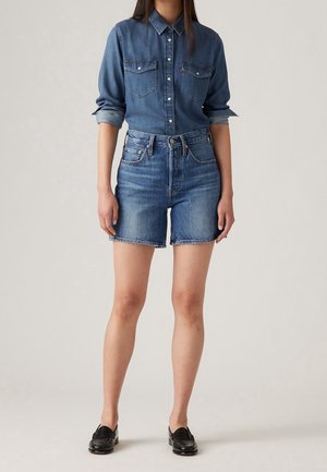 Femme portant une chemise en denim bleu à boutons avec manches retroussées, un short en denim bleu taille haute et des mocassins en cuir noirs, debout sur un fond uni.