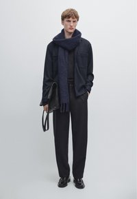 Young man wearing dark loose pants, black shirt, navy scarf, holding black leather bag, standing against plain light gray background.