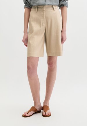 Beżowe szorty z wysokim stanem do kolan noszone z podwiniętą jasnoszarą koszulą i brązowymi płaskimi sandałami na białym tle.