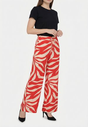 Donna con top nero a maniche corte, pantaloni larghi a stampa zebra rossi e beige, e scarpe nere con tacchi alti, in piedi con le mani giunte.