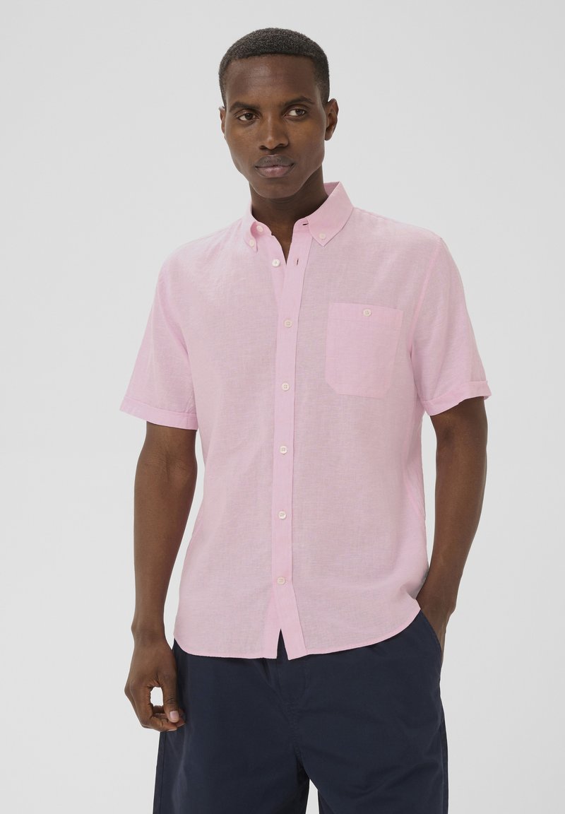 Jeune homme portant une chemise rose pâle à manches courtes avec poche avant et un short bleu marine foncé, debout avec une main dans la poche.