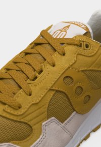 Κίτρινο και μπεζ αθλητικό παπούτσι με υφασμάτινες κορδόνια, πάνελ από δίχτυ, σουέντ επενδύσεις και λογότυπο της μάρκας στο πλάι και στη γλώσσα.