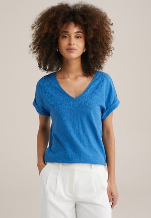 Korte mouw blauwe V-hals shirt met een gestructureerd luipaardpatroon. Stof lijkt zacht. Gecombineerd met witte, geplooide broek.