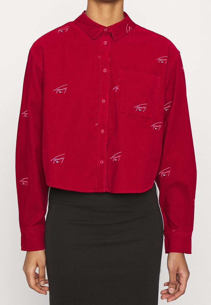 Red corduroy button-up shirt with long sleeves, featuring white embroidered logo details and a chest pocket. Cropped design.