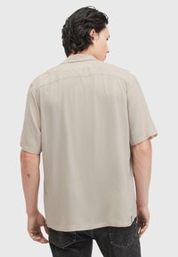 Camisa de manga corta en beige claro, textura suave, corte relajado, detalle en el yugo trasero, aberturas laterales, combinada con jeans oscuros.