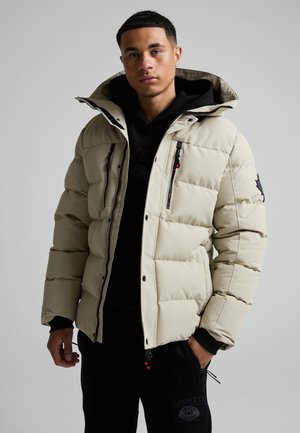 Ung mand iført en beige dunjakke over en sort hættetrøje og sorte bukser, stående mod en ensartet lysegrå baggrund.