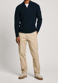 Dunkelblauer Strickpullover mit Reißverschluss am Kragen, kombiniert mit hellbeigen Chinos und beigefarbenen Sneakern. Einfaches, strukturiertes Design, lässige Passform.