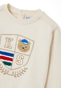 Felpa color crema con colletto rotondo. Presenta una grafica di un orso con un cappello blu, le lettere 'K' e 'S', e accenti colorati a righe.