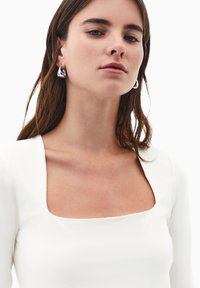 Donna con lunghi capelli castani che indossa orecchini a cerchio argento e una maglietta bianca a scollo quadrato su uno sfondo bianco uniforme.