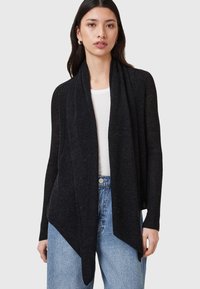 Black ribbed cardigan featuring an open front, long sleeves, and a pointed hem. Worn over a white top with relaxed-fit denim jeans.