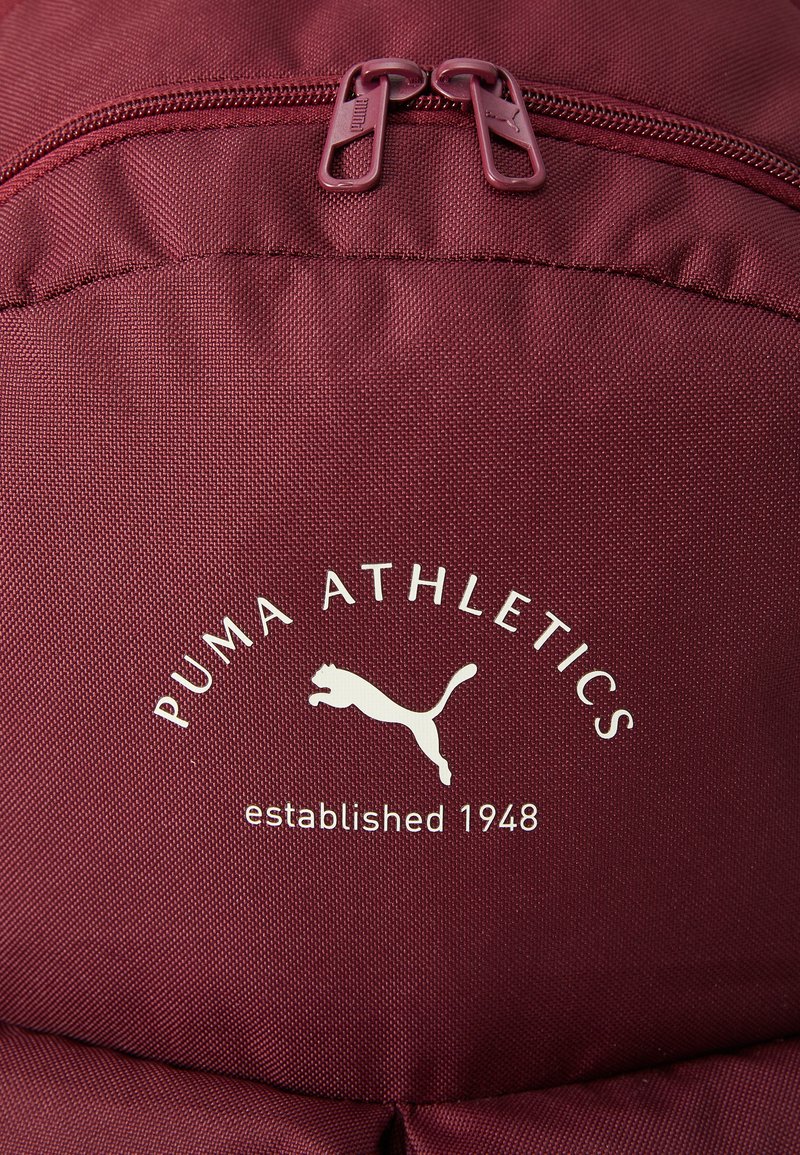 Weinrotfarbene Rucksack mit einem Reißverschlussfach, strukturiertem Stoff und einem weißen "PUMA ATHLETICS"-Logo mit einem springenden Puma-Grafik.