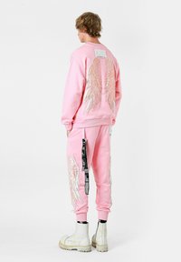 Sudadera rosa y pantalones a juego con bordado de alas metálicas, bolsillo lateral con detalle de correa negra, usados con botas blancas de tobillo.