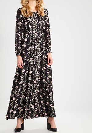 Nainen, jolla on pitkähihainen, pitkä musta mekko, jossa on vaaleanpunaiset kukkakuvioinnit ja vyötärö korostettu, yhdistettynä mustiin lohkopohjakenkiin.