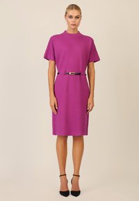Vestido corto fucsia de manga corta con un cuello alto, ceñido en la cintura con una correa negra y hebilla dorada, a la altura de la rodilla, de textura suave.