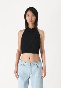 Svart halterneck crop top med drapert frontdesign. Laget av myk stoff, har høy halslinje og subtil tekstur. Kombinert med lyseblå jeans.