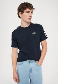 Tamsiai mėlynas trumpomis rankovėmis marškinėliai su apvalia iškirpte, turintys logotipo siuvinėjimą ir baltas šonines juostas. Pagaminti iš lygaus audinio,uriantys slim fit dizainą.