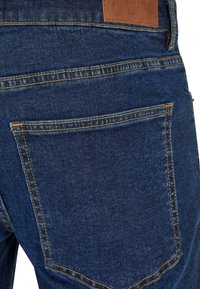 Jean en denim bleu foncé avec des coutures jaunes, un patch en cuir sur la taille et deux poches arrière avec des coutures arrondies.