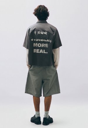 Person mit lockigem Haar steht mit dem Rücken zur Kamera, trägt ein schwarzes Shirt mit dem Schriftzug "Less visible, more real,", karierte Shorts, weiße Socken und schwarze Sneakers vor einfachem Hintergrund.