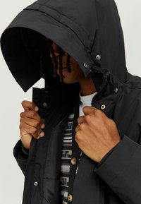 Black padded jacket with a hood, featuring snap buttons and a zip closure. Underneath, a patterned sweater is partially visible.