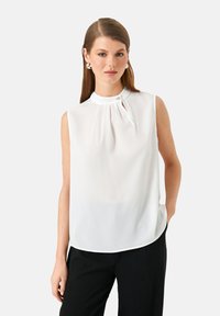 Junge Frau mit glattem braunem Haar, die eine weiße ärmellose Bluse mit hochgeschlossenem Kragen und schwarze Hosen trägt, steht vor einem weißen Hintergrund.