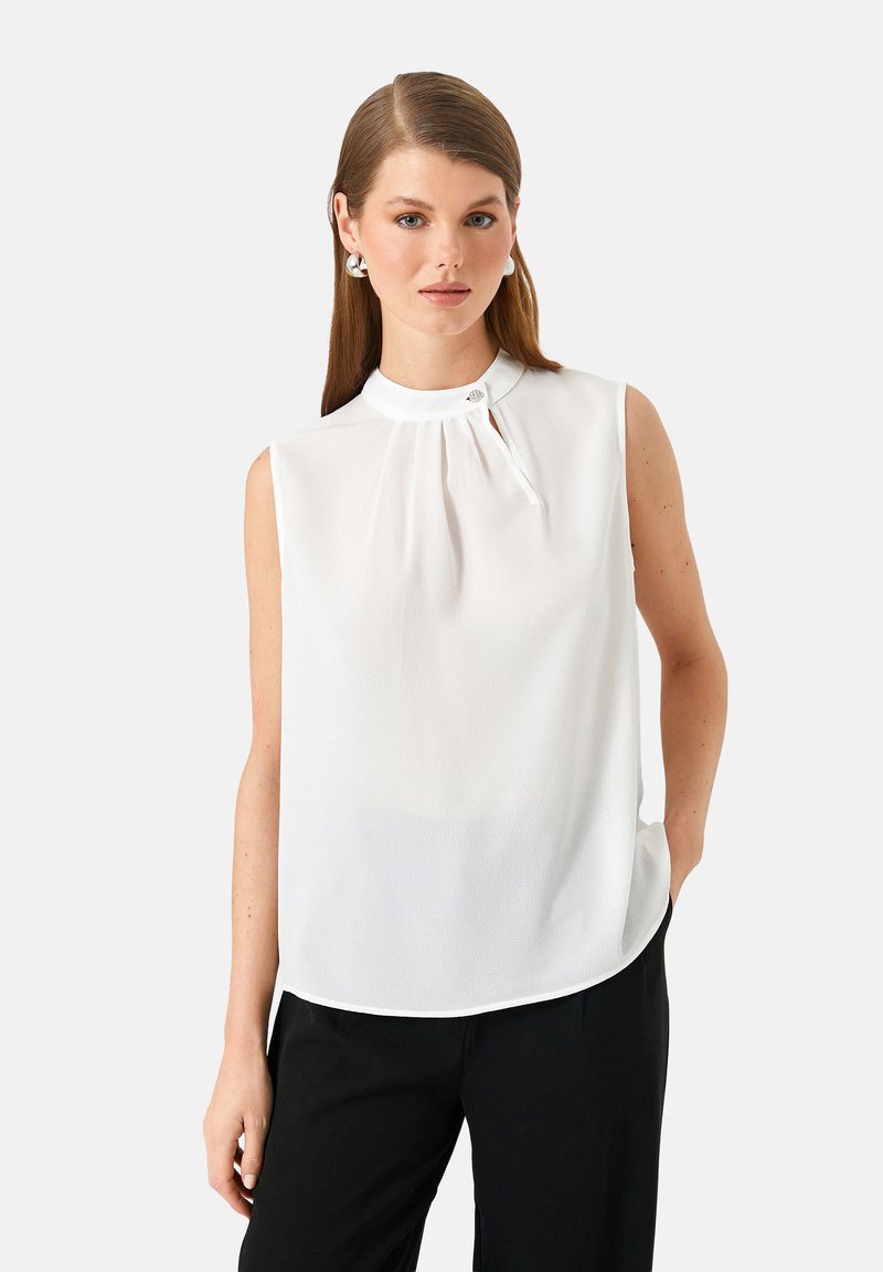 Junge Frau mit glattem braunem Haar, die eine weiße ärmellose Bluse mit hochgeschlossenem Kragen und schwarze Hosen trägt, steht vor einem weißen Hintergrund.