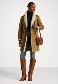 Cappotto in shearling marrone con fodera crema, abbinato a un dolcevita blu, jeans in denim e stivali marroni fino al ginocchio. Gli accessori includono una borsa a tracolla marrone.
